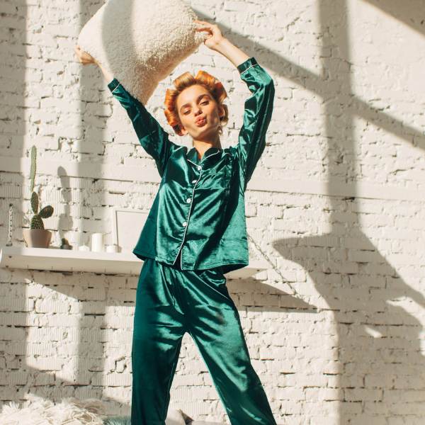 Woman in satin pajamas holding pillow playfully in a sunlit bedroom